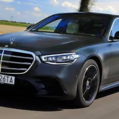 Elegancki, ciemnoszary Mercedes S580e sunie po drodze wśród zielonych pól i drzew.