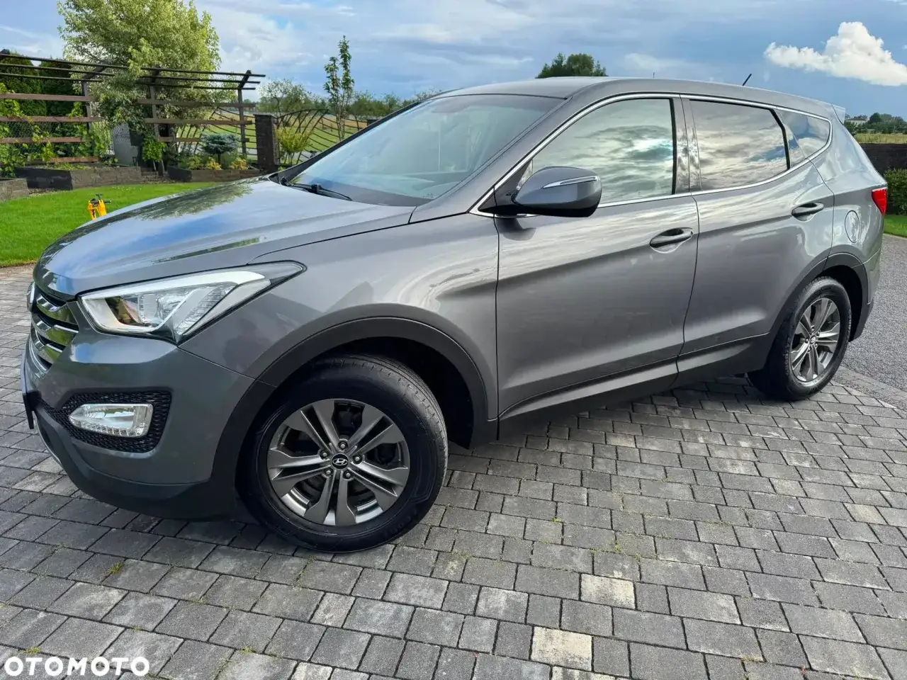 Szary Hyundai Santa Fe zaparkowany na podjeździe z kostki brukowej, w tle ogród i błękitne niebo.